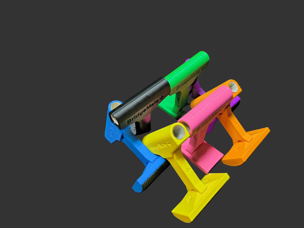 Bridge Mate Plus 3D-printed elevated pool cue bridge heads in black, green, blue, pink, yellow, and orange — stacked on LH Inventions carry bag showing BridgeMate+ branding