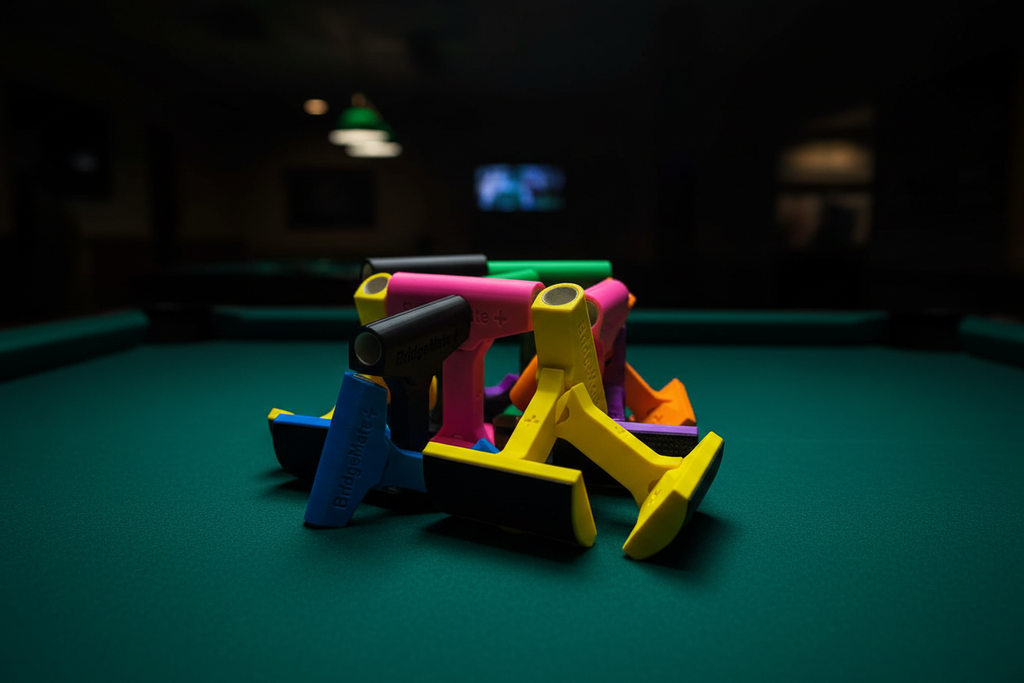 Bridge Mate Plus 3D-printed elevated billiard bridge heads in multiple colors including yellow, pink, black, blue, green, and orange — displayed on green pool table felt in dark billiard room