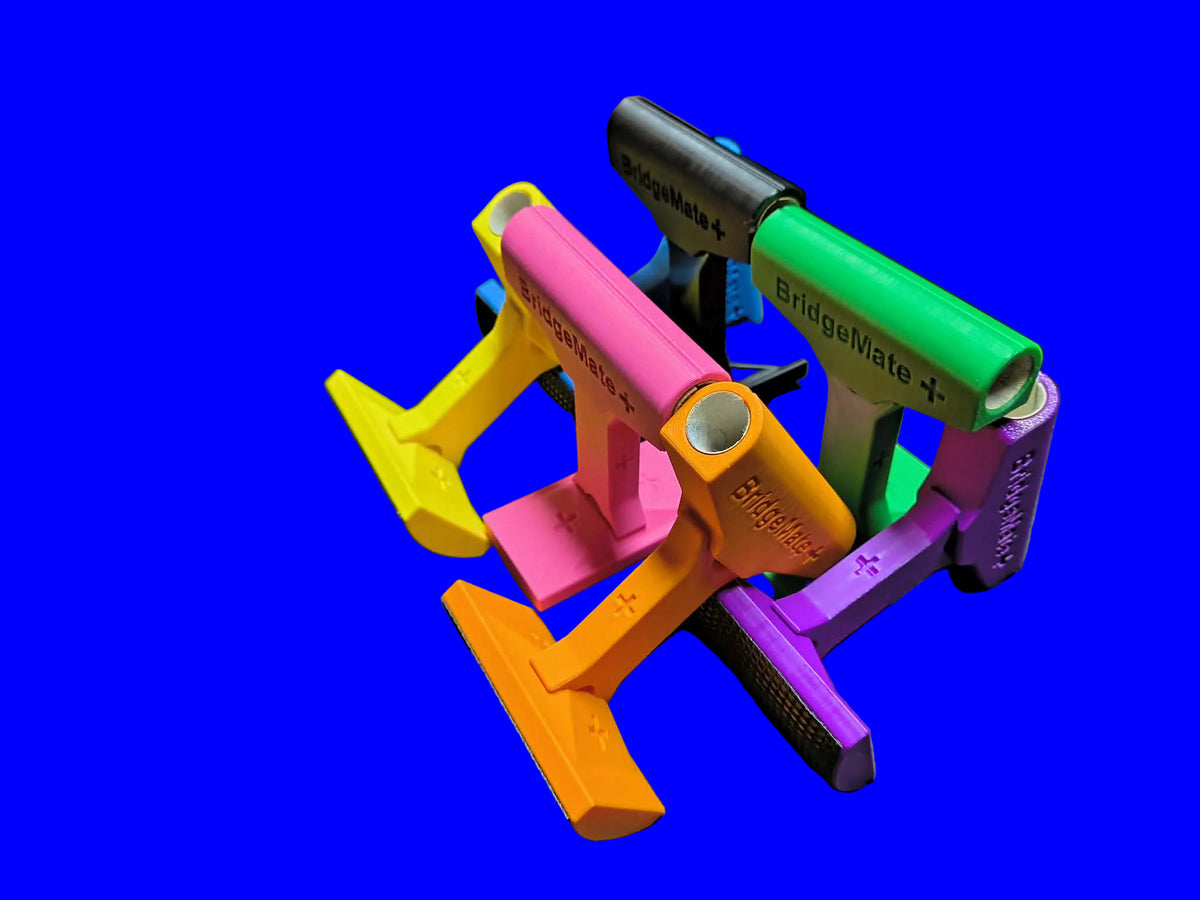 Bridge Mate Plus 3D-printed elevated billiard bridge heads in yellow, pink, green, black, orange, and purple — fanned out on LH Inventions carry bag showing elevated arch and cue ferrule opening